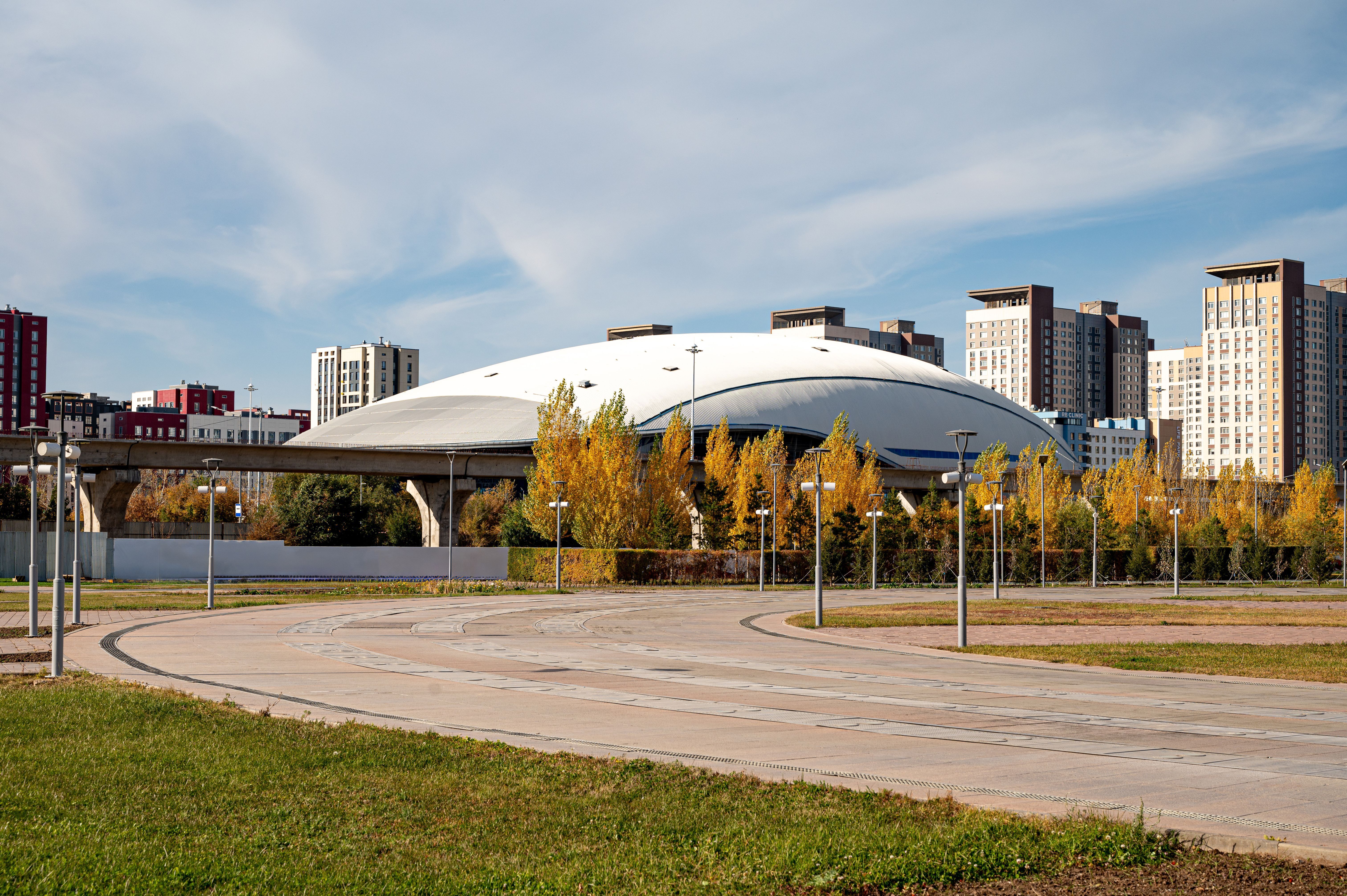 The view of the Saryarka Velodrome building, a modern sports complex in Astana, Kazakhstan, is framed by autumn trees and residential buildings against a clear sky