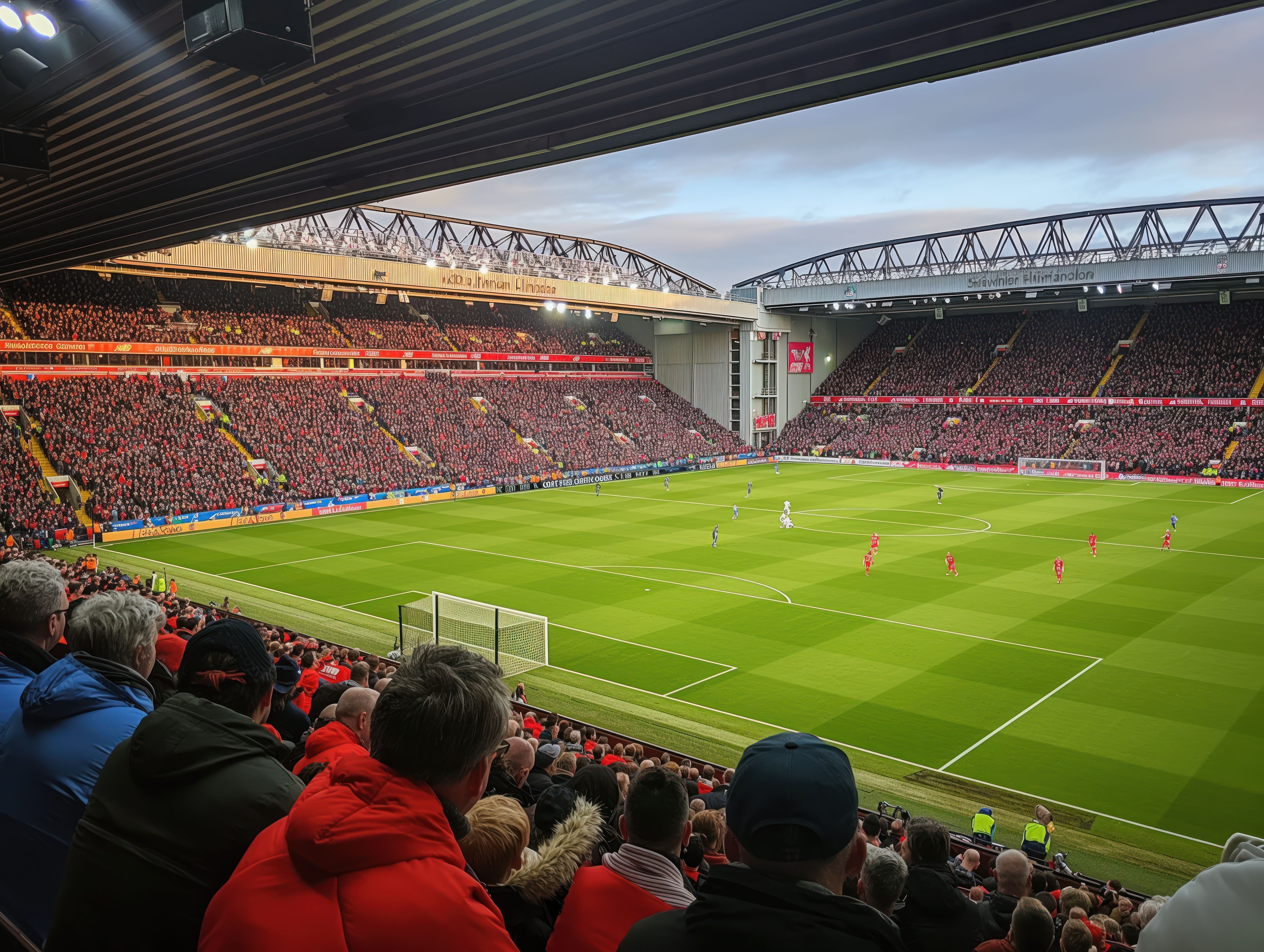 Crowd of people gathered at Anfield Stadium watching a football game. Green grass and grandstands fill the frame. Excited fans cheering and waving their arms in the stands. Sports event atmosphere.