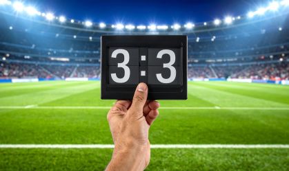 A hand holding a scoreboard displaying a 3-3 tie during a soccer game in a stadium.